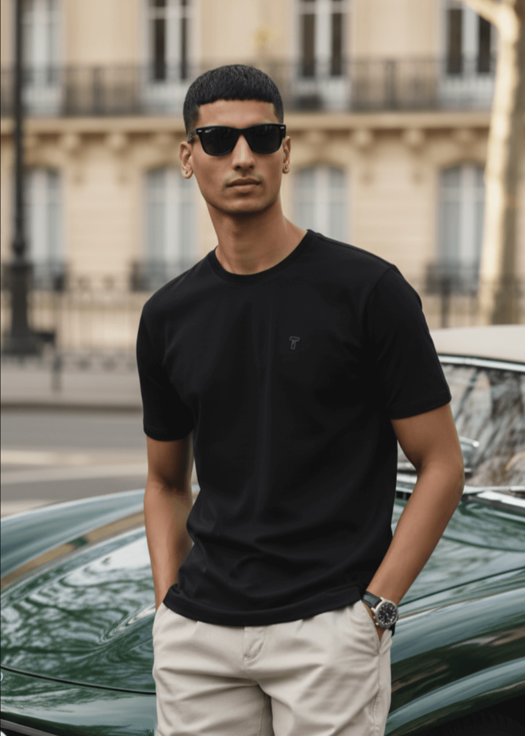 Man wearing a black t-shirt and sunglasses standing in front of a car with a blurred building in the background